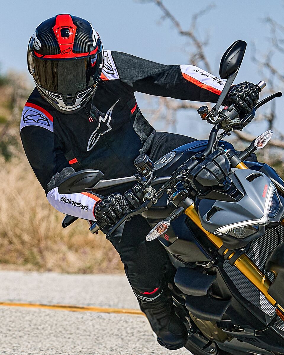 Motociclista in piega su una moto naked sportiva, indossa la giacca Alpinestars T-SPS Air v2 in nero con accenti bianchi e rosso fluo, abbinata a un casco nero e rosso e guanti neri, abbigliamento tecnico in tessuto orientato alle prestazioni per la guida sportiva urbana.