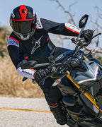 Motociclista in piega su una moto naked sportiva, indossa la giacca Alpinestars T-SPS Air v2 in nero con accenti bianchi e rosso fluo, abbinata a un casco nero e rosso e guanti neri, abbigliamento tecnico in tessuto orientato alle prestazioni per la guida sportiva urbana.