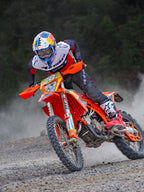 Pilota di motocross in azione con maglia Alpinestars Pro-Dura in nero, rosso acceso e bianco, alla guida di una moto da cross arancione su un sentiero fuoristrada polveroso, abbinata a pantaloni neri e stivali rossi e bianchi