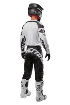 Alpinestars 2024 Racer Hana Pants, pantaloni da motocross, nero e bianco con loghi sulla parte posteriore, vista posteriore, indossati con maglia bianca coordinata, casco nero e stivali bianchi