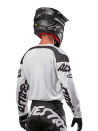 Alpinestars Maglia Racer Hana 2024, bianco e nero, vista posteriore che mostra la vestibilità ergonomica e il branding evidente sulle maniche, abbinata a un casco da motocross nero e pantaloni coordinati bianchi e neri, equipaggiamento da gara ad alte prestazioni per il fuoristrada