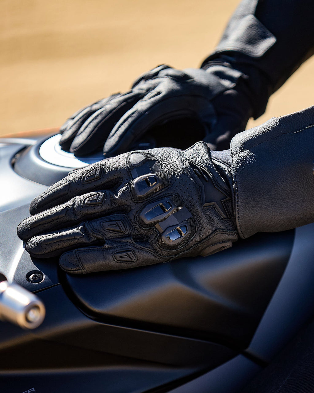 Guanti Alpinestars SP X 7, neri, guanti da moto a polsino corto in pelle perforata, mani appoggiate sul serbatoio di una moto, con protezione nocche in TPU a doppia densità e logo in rilievo, progettati per la guida sportiva e stradale.