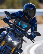 Motociclista in piega in curva che indossa la giacca Alpinestars T-Stunt Air in nero, blu scuro e blu brillante, abbinata a casco e guanti neri, scatto d'azione su strada asfaltata, abbigliamento tecnico sportivo da moto