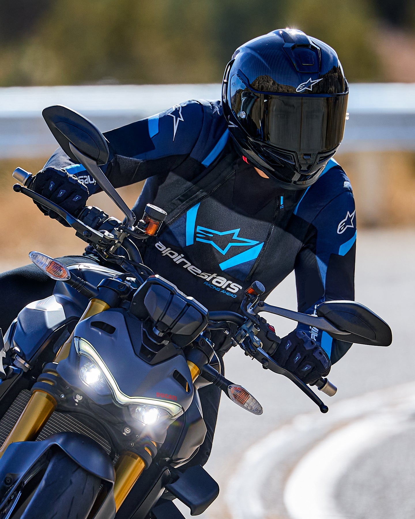 Motociclista in piega in curva che indossa la giacca Alpinestars T-Stunt Air in nero, blu scuro e blu brillante, abbinata a casco e guanti neri, scatto d'azione su strada asfaltata, abbigliamento tecnico sportivo da moto