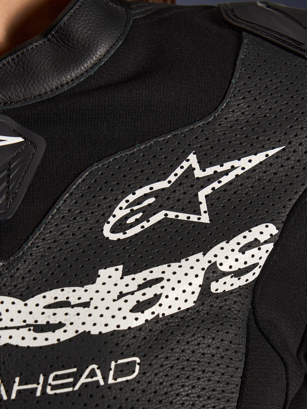 Primo piano della tuta in pelle Alpinestars Stella GP-R7 2 Pezzi in nero, caratterizzata da pannelli in pelle traforata, un grande logo Alpinestars bianco sul petto e sezioni in tessuto elastico traspirante.