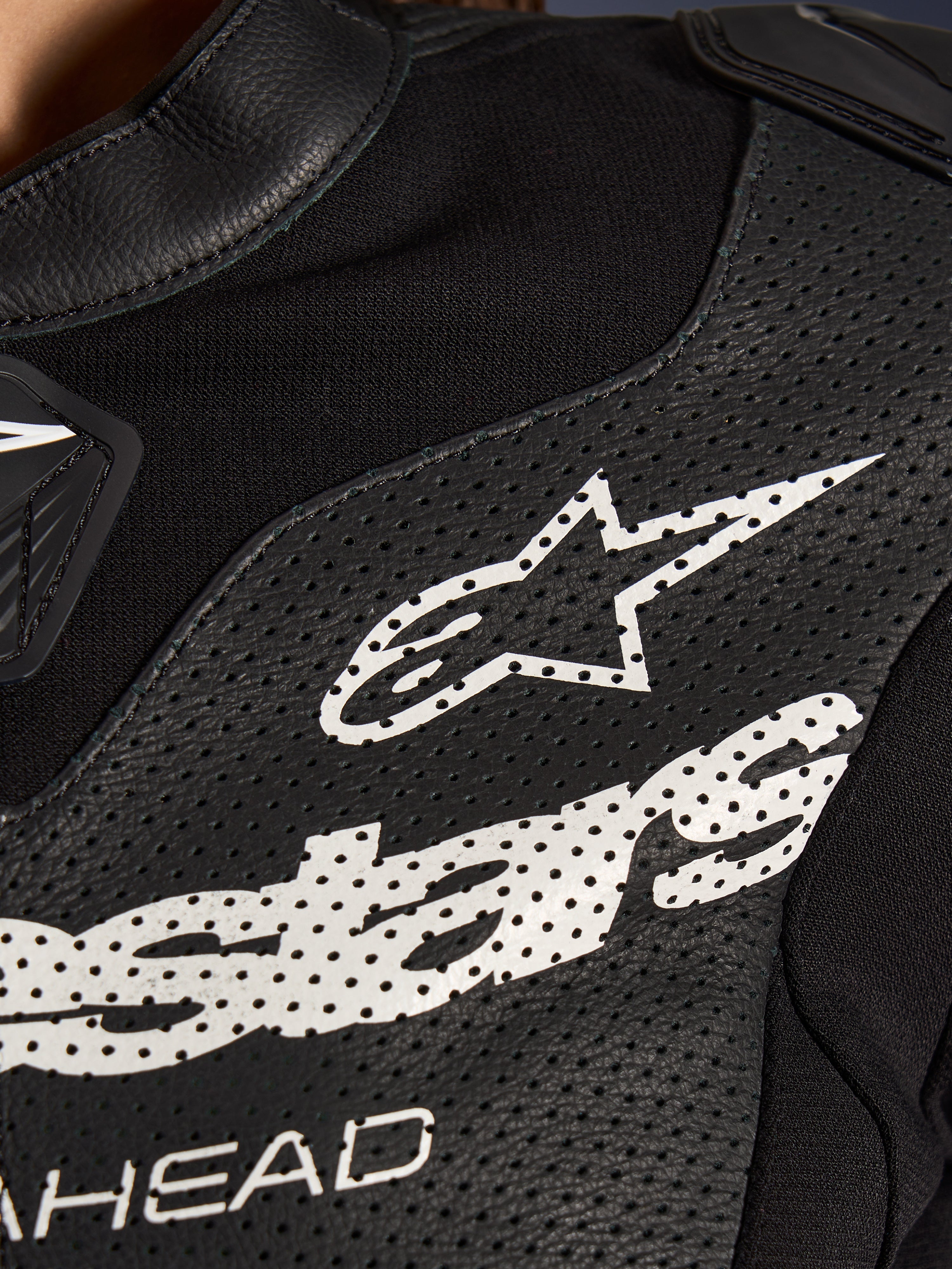 Primo piano della tuta in pelle Alpinestars Stella GP-R7 2 Pezzi in nero, caratterizzata da pannelli in pelle traforata, un grande logo Alpinestars bianco sul petto e sezioni in tessuto elastico traspirante.