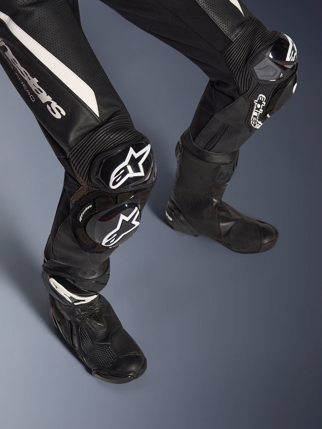 Dettaglio delle gambe della tuta in pelle Alpinestars Stella GP-R7 2 Pezzi, nera con accenti bianchi, con protezioni ginocchia e slider GP DFS RR con marchio Alpinestars, abbinata a stivali da corsa neri, progettata per prestazioni in pista