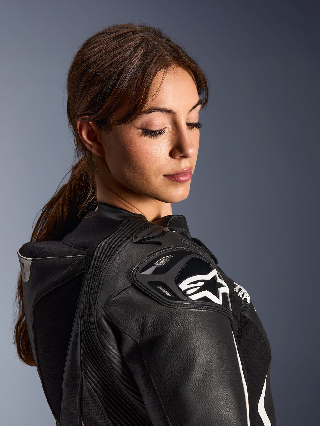 Donna che indossa la tuta in pelle Alpinestars Stella GP-R7 2 pezzi, primo piano della spalla in pelle nera con protezione DFS RR e logo a stella bianco, gobba aerodinamica integrata e dettagli traforati