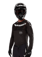 Alpinestars 2024 Supertech Ward Jersey, maglia da motocross, nero e bianco, abbigliamento tecnico a maniche lunghe con branding Alpinestars in evidenza, indossata con casco e guanti coordinati, vestibilità atletica per la guida fuoristrada