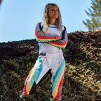 Pilota di motocross donna con pantaloni Alpinestars Stella Techstar 2024 e maglia coordinata, design Black Multicolor con base bianca e strisce blu, gialle e rosse, in piedi con le braccia incrociate su pista sterrata, abbigliamento professionale da gara fuoristrada