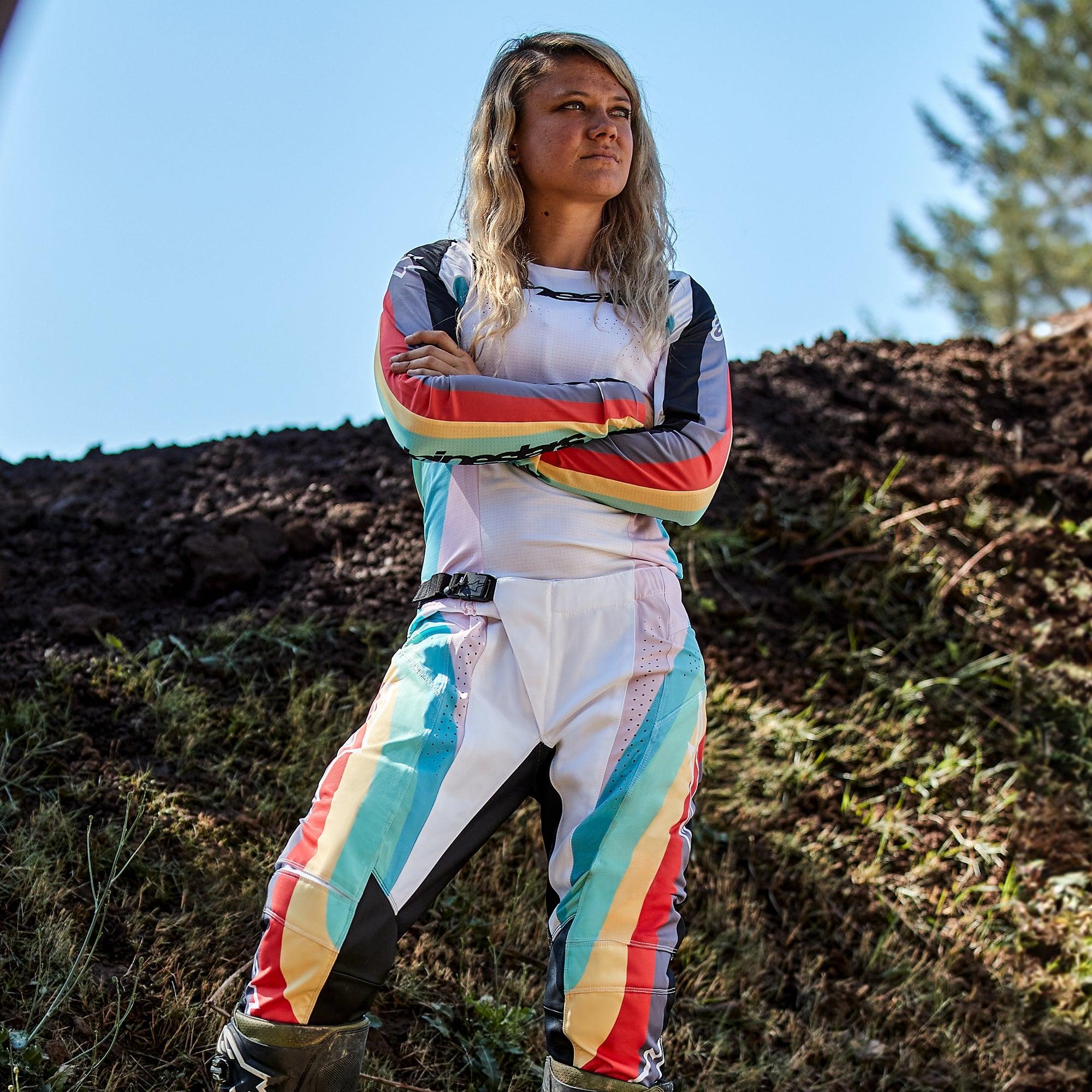 Pilota di motocross donna con pantaloni Alpinestars Stella Techstar 2024 e maglia coordinata, design Black Multicolor con base bianca e strisce blu, gialle e rosse, in piedi con le braccia incrociate su pista sterrata, abbigliamento professionale da gara fuoristrada