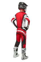 Alpinestars 2024 Techstar Ocuri Pants, pantaloni da motocross, rosso Mars, bianco e nero, vista posteriore di un pilota di motocross con attrezzatura da gara completa, inclusa maglia abbinata, casco nero e stivali bianchi e neri, mette in risalto il marchio Alpinestars e la struttura resistente.