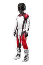 Pilota di motocross con abbigliamento completo Alpinestars, pantaloni Techstar Ocuri 2024 in Mars Red White Black, maglia coordinata, casco nero con occhiali, guanti neri e bianchi, stivali da motocross bianchi, completo professionale da corsa off-road