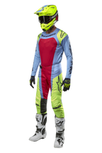 Pilota di motocross con pantaloni Alpinestars Techstar Ocuri 2024 in azzurro, giallo fluo e rosso, abbigliamento da gara coordinato con maglia abbinata, casco giallo fluo e stivali bianchi, vista frontale del completo da competizione fuoristrada