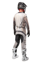 Alpinestars Techstar Pneuma Pants 2024, pantaloni da motocross, sabbia scura e grigio, vista posteriore indossata da un pilota con maglia coordinata, stivali bianchi e neri e casco nero, caratterizzati da vistoso marchio Alpinestars sulla seduta e pannelli scuri rinforzati