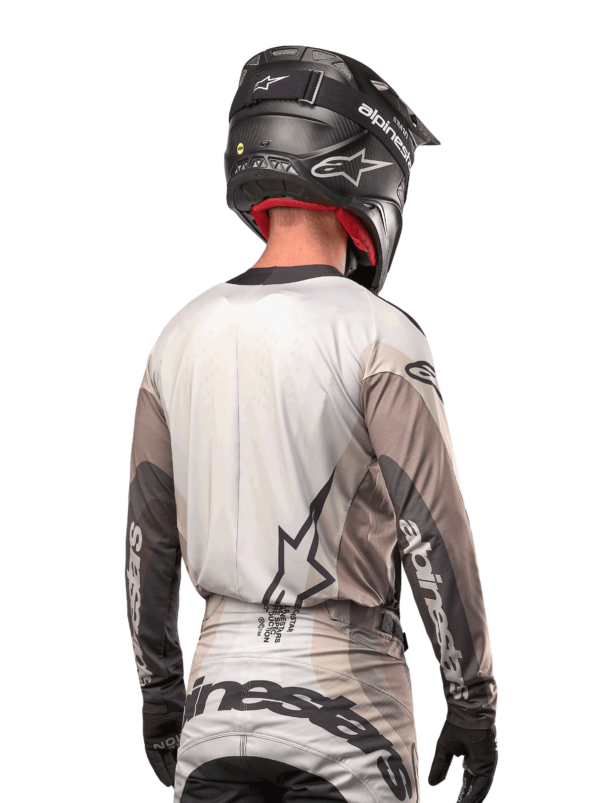 Pilota di motocross visto da dietro che indossa la maglia Alpinestars 2024 Techstar Pneuma in Dark Sand Iron Dust Gray, abbinata a un casco nero con finitura in carbonio, con un grande logo a stella sulla schiena e marchio Alpinestars sulle maniche.