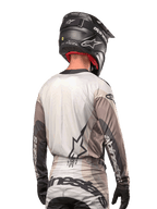 Pilota di motocross visto da dietro che indossa la maglia Alpinestars 2024 Techstar Pneuma in Dark Sand Iron Dust Gray, abbinata a un casco nero con finitura in carbonio, con un grande logo a stella sulla schiena e marchio Alpinestars sulle maniche.