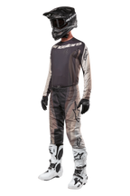Pantaloni Alpinestars Techstar Pneuma 2024, pantaloni da motocross, Dark Sand Iron Dust Gray, indossati come parte di un set completo con maglia coordinata, casco nero, guanti neri e stivali bianchi, caratterizzati da motivi mimetici desertici tonali con grandi loghi.