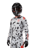 Alpinestars 2024 Techstar Rantera Jersey, maglia da motocross, Haze Gray Camo con motivi astratti neri e accenti rossi, design a maniche lunghe indossato con casco e guanti neri coordinati, abbigliamento tecnico da competizione off-road