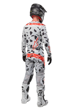 Pilota di motocross visto da dietro, indossa pantaloni Alpinestars 2024 Techstar Rantera, Haze Gray Camo con macchie nere e loghi rossi, maglia coordinata, casco nero e stivali bianchi, completo da gara fuoristrada