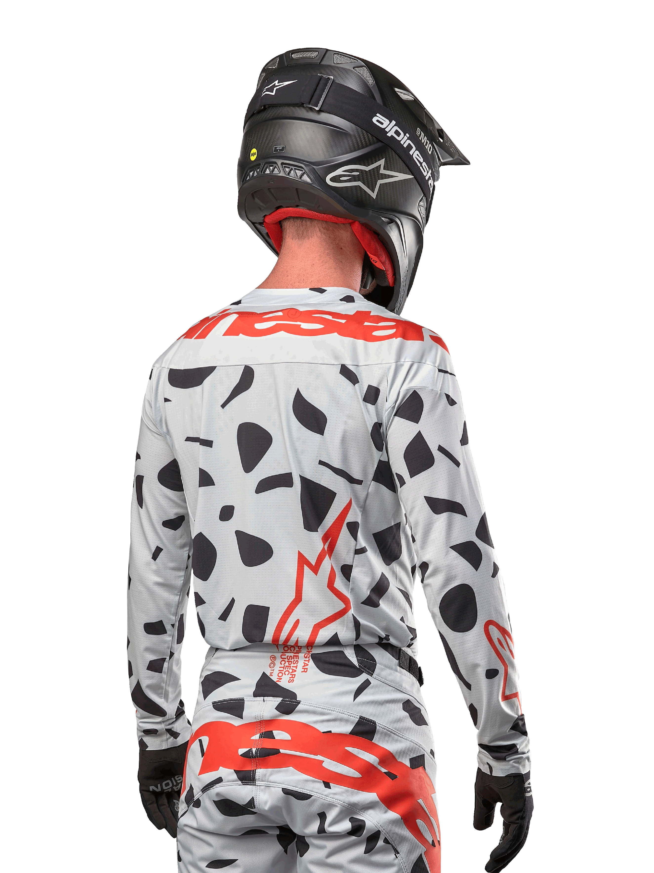 Pilota di motocross visto di spalle che indossa la maglia Alpinestars 2024 Techstar Rantera in Haze Gray Camo, con base grigio chiaro e macchie grafiche nere, vivaci loghi Alpinestars rossi su schiena e spalle, abbinata a pantaloni coordinati e casco nero.