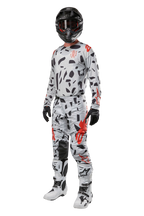 Pantaloni Alpinestars Techstar Rantera 2024, pantaloni da motocross, Haze Gray Camo, indossati con maglia coordinata, casco nero e stivali bianchi e neri, con motivi astratti neri e accenti rossi, equipaggiamento completo da gara fuoristrada
