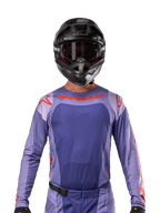 Alpinestars Techstar Ocuri Jersey 2024, maglia da motocross viola e arancione acceso, indossata da un pilota con casco e occhiali neri, dotata di perforazioni tagliate al laser sul petto, vestibilità atletica race fit e loghi rossi su spalle e petto.