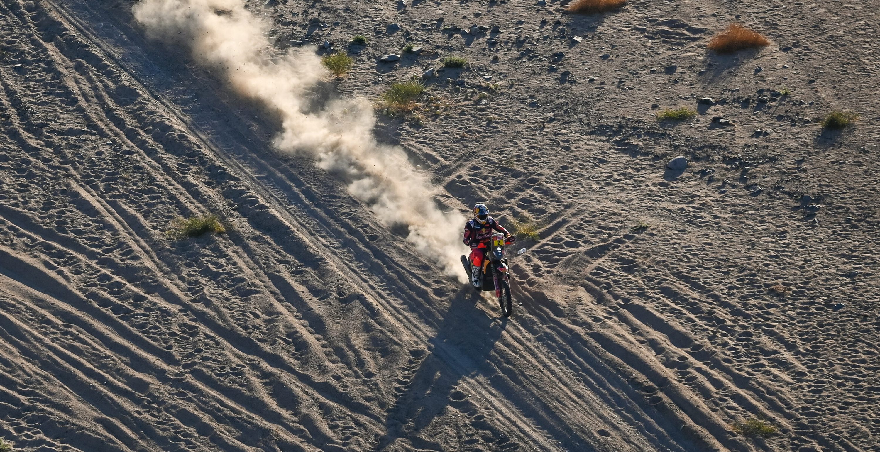 Toby Price riding a Dakar rally bike on a sandy desert landscape with dust trail