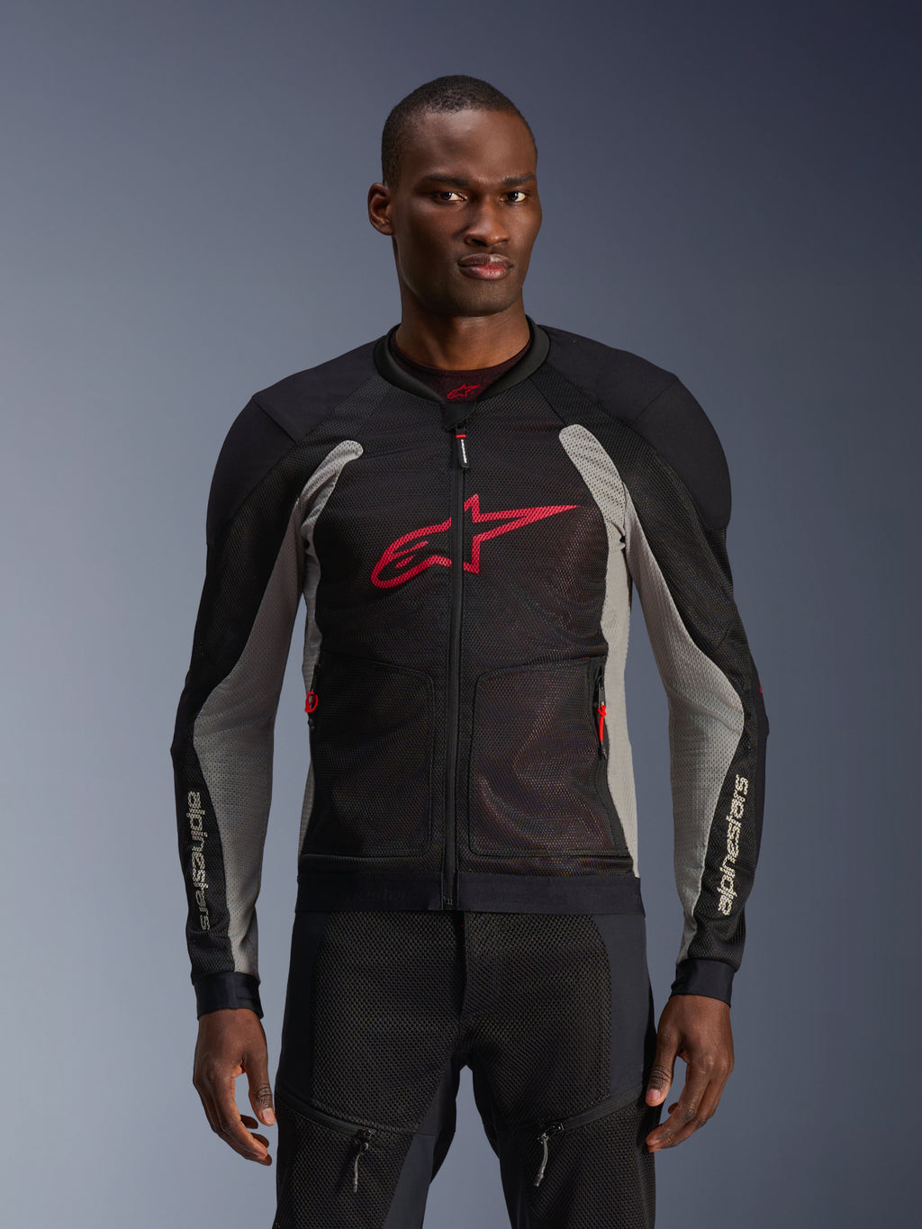 Giacca Alpinestars Troop-Air, giacca da moto in mesh, nero e grigio cenere con accenti rosso acceso, indossata da un modello, dotata di pannelli in mesh tecnico 2D traspirante per la ventilazione, grande logo Astars rosso sul petto e design tecnico sportivo per la guida urbana