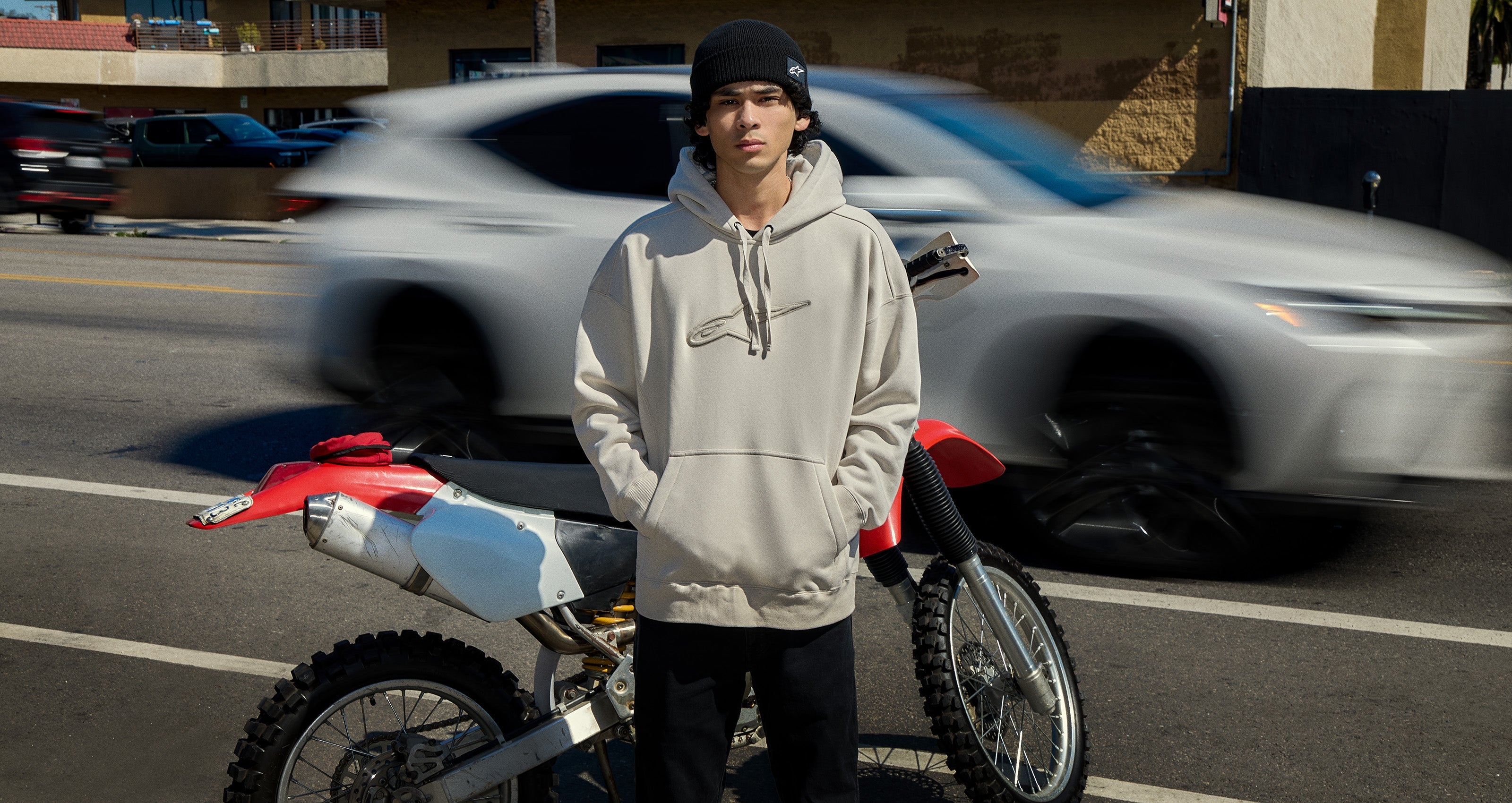 Alpinestars Emergent Hoodie, lifestyle pullover, Light Beige, with embroidered logo, worn by a man next to a dirt bike.