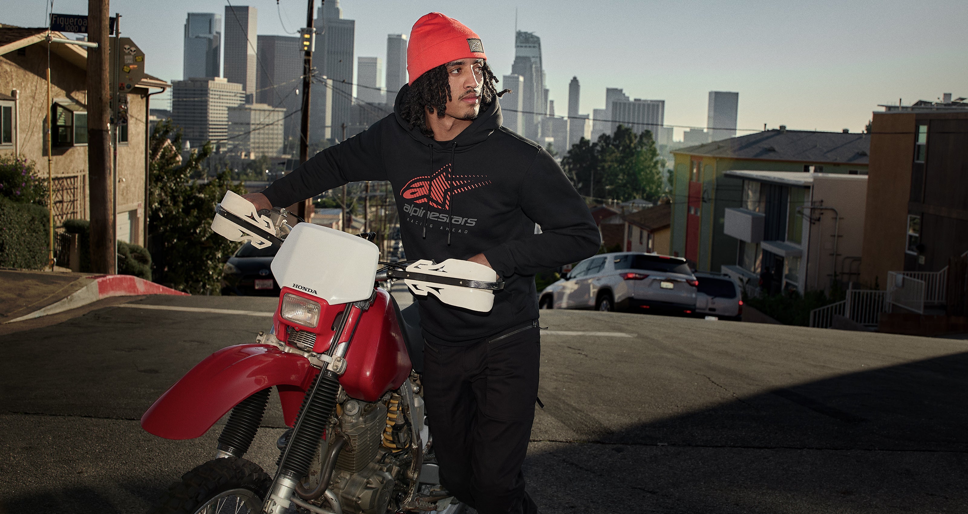 Alpinestars red beanie and black logo hoodie, urban lifestyle apparel worn by a man on a motorcycle with a city skyline.
