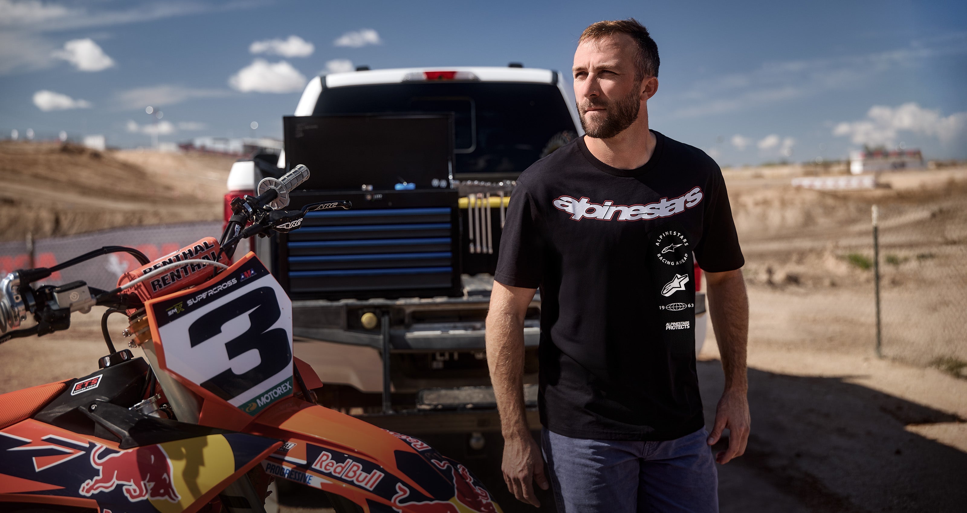 Alpinestars black sportswear t-shirt with white graphic logos, worn by a man at a motocross track next to a racing bike.