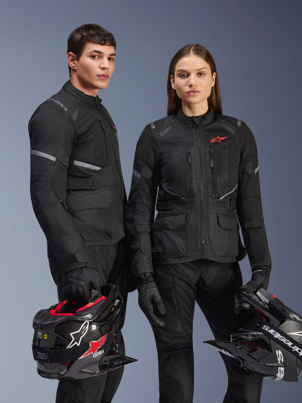 Modello e modella che indossano giacche Alpinestars Stella Andes Air Drystar® in nero, abbigliamento tecnico per mototurismo adventure con loghi a stella rossi, pannelli di ventilazione in rete e costruzione in tessuto tecnico, modelli con caschi adventure in uno scatto in studio