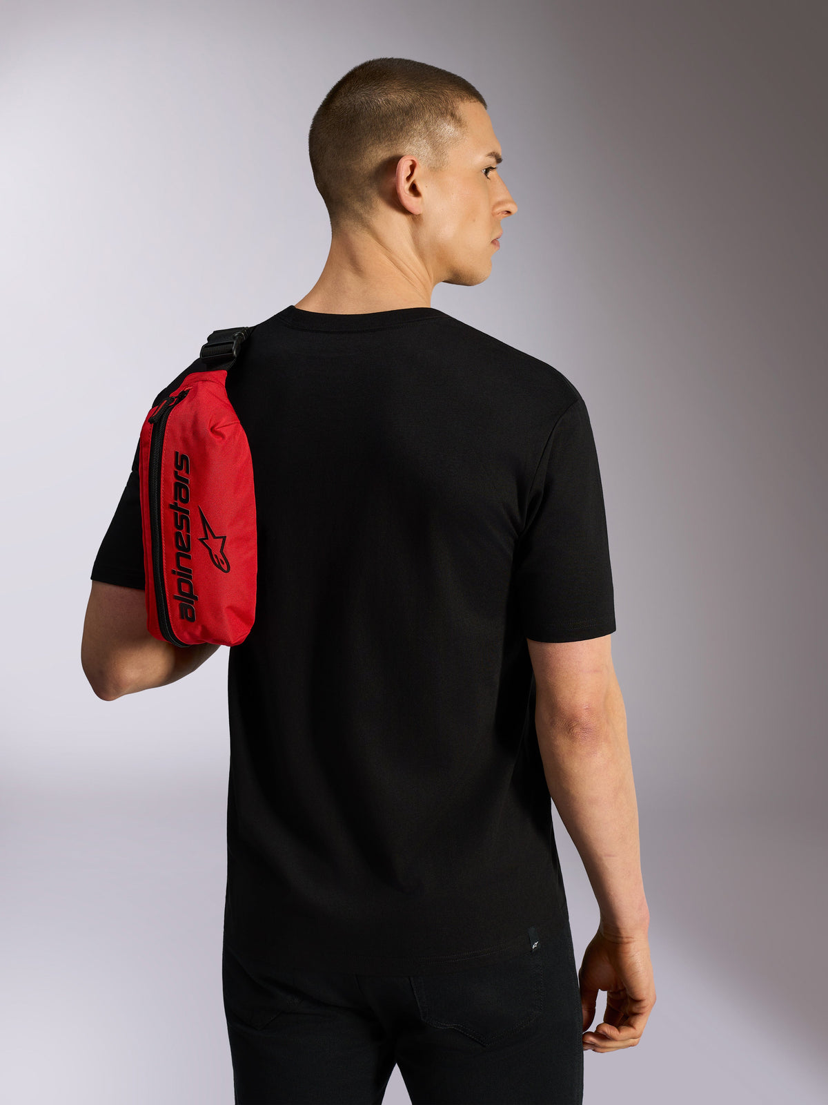 Alpinestars Only Linear Hip Bag, rosso e nero, indossato a tracolla da un modello in t-shirt nera, con cerniera verticale e marchio Alpinestars nero, accessorio urbano resistente.
